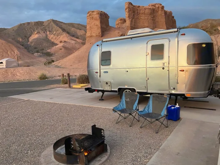 This Airstream RV rental is perfectly set up for a desert camp, featuring a cozy fire pit and chairs by scenic mountains.. Airstream Bambi 2023