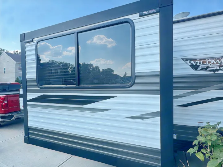 Modern rv rental travel trailer side: striped exterior, large window reflecting blue sky, clouds. Forest River Wildwood.. Forest River Wildwood FSX 2023