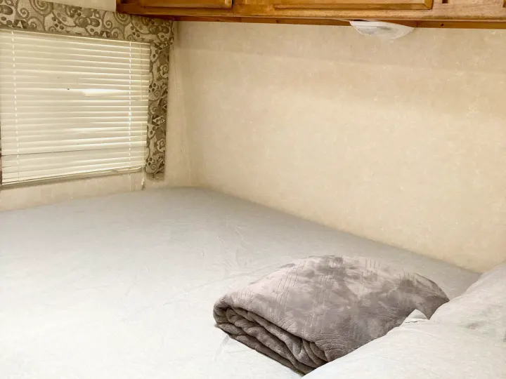 Cozy RV rental bedroom featuring a comfortable bed with light grey bedding, a folded blanket, and window with blinds.. Rage'N Toy Hauler 2006