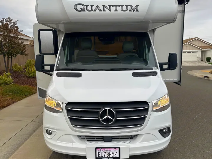 Frontal view of a white Mercedes-Benz Quantum RV rental from Camping World, parked in a residential setting with lights on.. Thor Quantum 2023