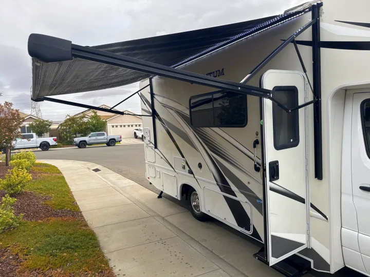 This RV rental, a Class C motorhome, features an extended awning with bright LED lights, parked on a residential street.. Thor Quantum 2023