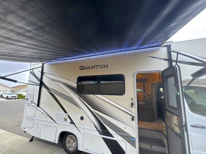 A modern RV rental, featuring an extended awning with bright LED lights, door open, parked on a residential street.. Thor Quantum 2023