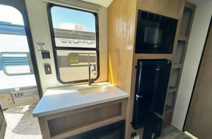 RV rental kitchen: counter with sink, microwave, & mini-fridge. Window view shows another 'Wolf Pup' RV outside.. Forest River Cherokee Wolf Den 2024