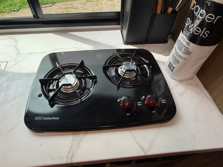Two-burner Suburban gas stove on a white countertop in an RV rental kitchen, with controls, paper towels, and window view.. Forest River Salem 2026