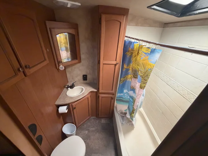 Interior of an RV rental bathroom, featuring a toilet, sink, mirror, and a shower/tub combo with a tropical curtain.. Crossroads RV Sunset Trail Super Lite 2015