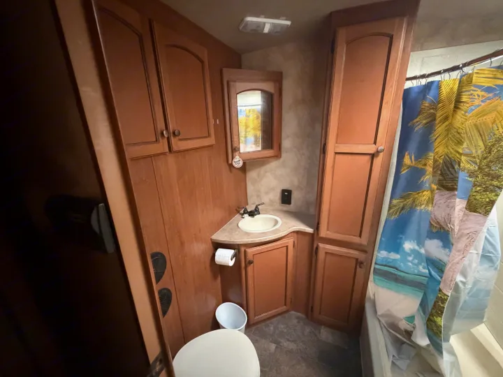 Compact RV rental bathroom: features a toilet, sink, wood cabinets, and a shower with a tropical palm tree curtain.. Crossroads RV Sunset Trail Super Lite 2015