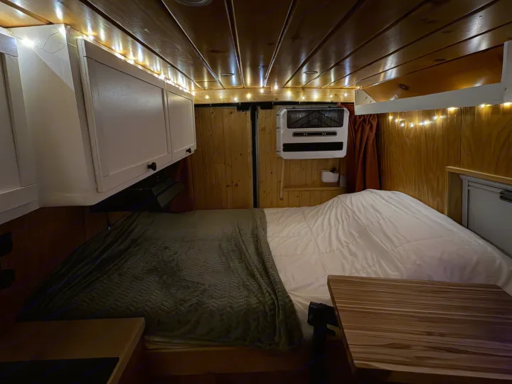 Cozy RV rental interior shows a bed, wooden walls, string lights, a/c unit, and overhead cabinets for a comfortable adventure.. Dodge Ram 2500 2024