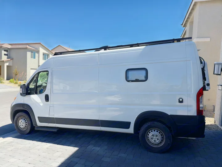 White Ram ProMaster camper van, parked on a paved driveway with a roof rack and side window. Ideal for rv rental adventures.. Dodge Ram 2500 2024