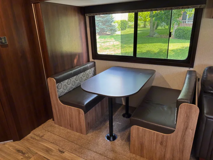 RV rental dinette: cozy interior with dark benches, patterned backrests, and a window overlooking a green yard. Ideal for meals.. Jayco Jay Feather 2018