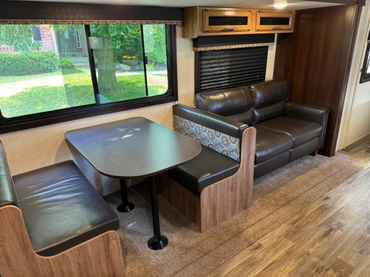 Inside an rv rental: a dark dinette with comfortable seating, a matching sofa, and windows overlooking a green yard.. Jayco Jay Feather 2018