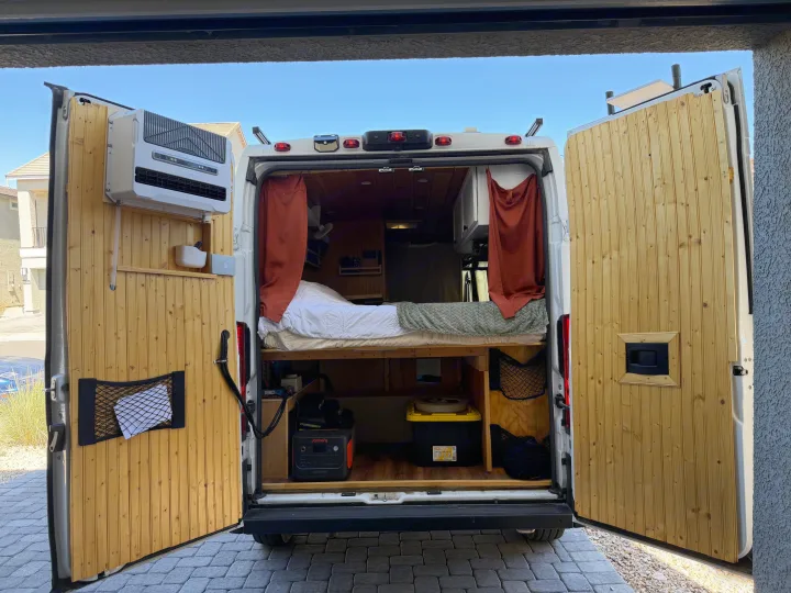Inside a custom rv rental camper van: rear doors open to wood-paneled interior, bed, storage, portable AC, and off-grid power setup.. Dodge Ram 2500 2024