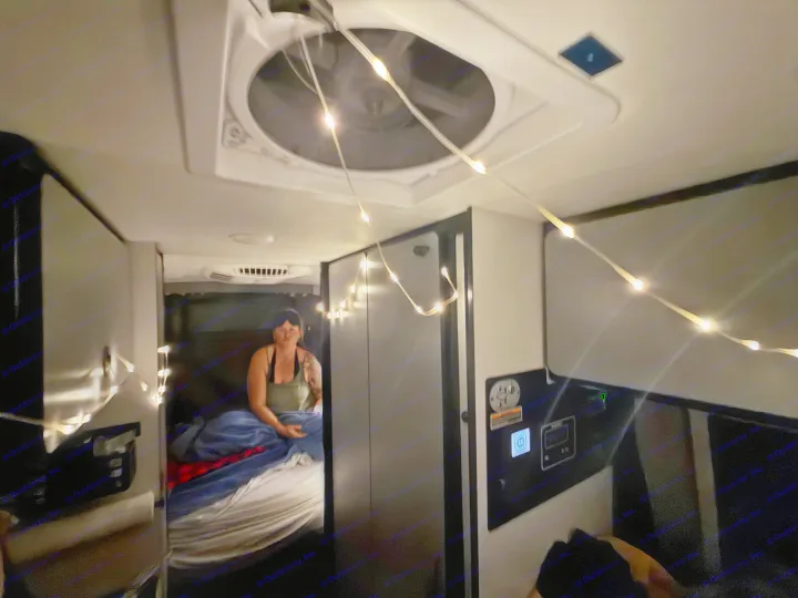 Cozy rv rental interior: a woman rests on a bed, lit by string lights. Ceiling fan & control panel visible for comfort.. Winnebago Revel 2025