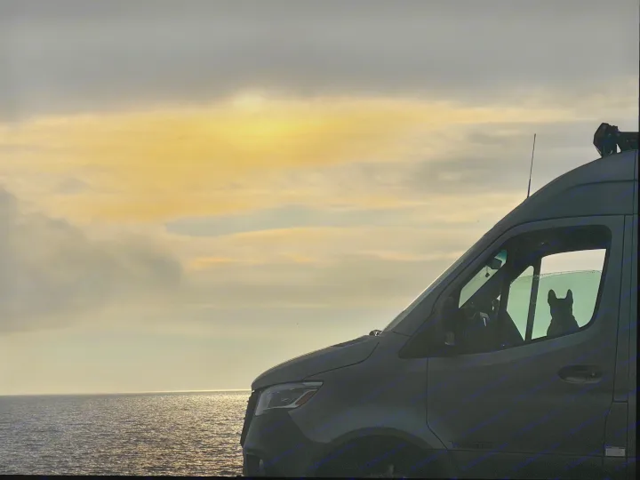 A dog's silhouette watches the ocean from an **rv rental** van at sunset, under a golden, cloudy sky.. Winnebago Revel 2025