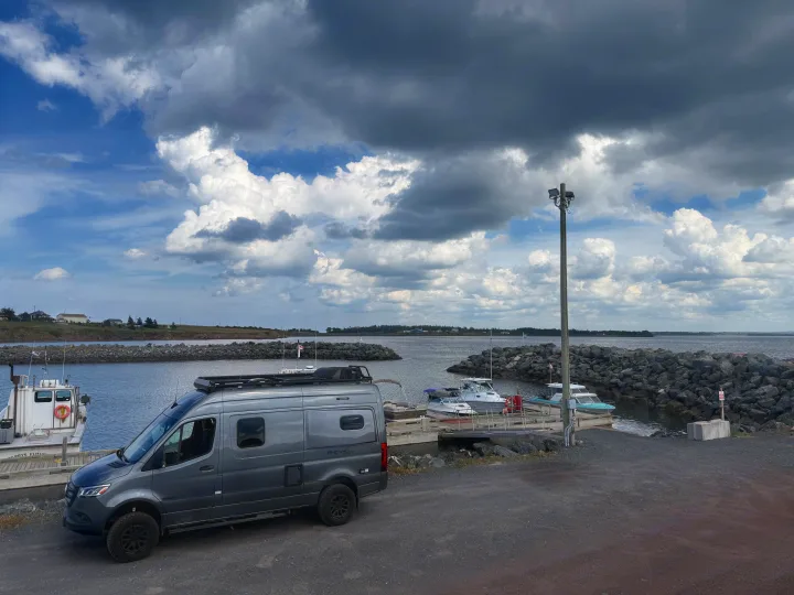 A grey Mercedes Revel RV rental camper van parked at a harbor with boats and a rocky shoreline under dramatic clouds.. Winnebago Revel 2025