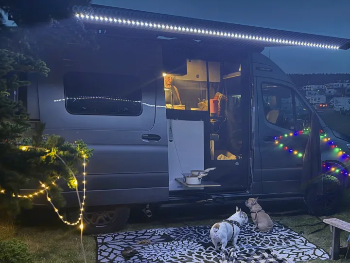 Cozy RV rental setup at night: a camper van with a person inside, two French Bulldogs, and festive outdoor lights.. Winnebago Revel 2025