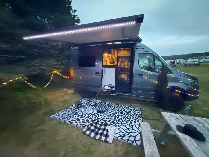Cozy **rv rental** campervan setup at an evening campsite, with illuminated awning, string lights, and a person inside.. Winnebago Revel 2025