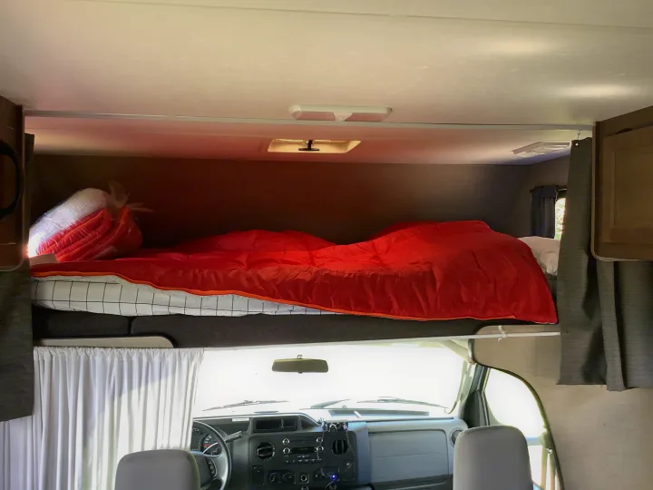 This RV rental interior showcases a cozy red bunk bed positioned above the driver's cockpit, visible with its dashboard.. Thor Majestic 2018