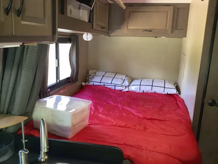 This RV rental bedroom features a comfy bed with a red comforter, patterned pillows, cabinets, and a small sink.. Thor Majestic 2018