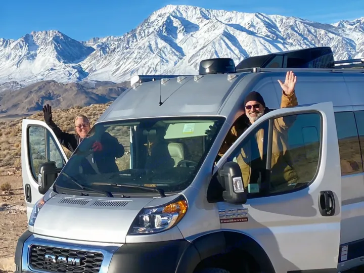 Happy couple waving from their RV rental van against a backdrop of stunning, snow-capped mountains.. Thor Sequence 2022