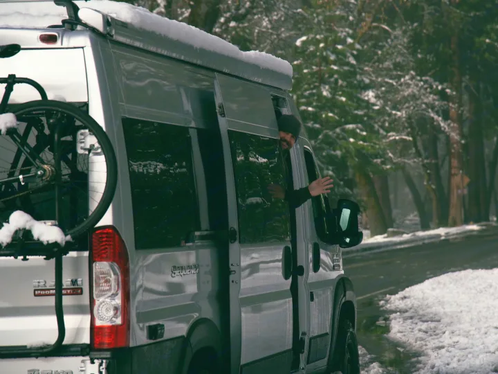 A person waves from an RV rental van, parked on a snowy road among trees. A bike is mounted on the rear.. Thor Sequence 2022