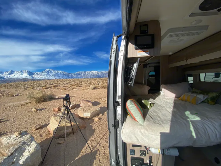 RV rental van interior with bed facing majestic snow-capped mountains in a desert landscape. A camera on tripod waits.. Thor Sequence 2022