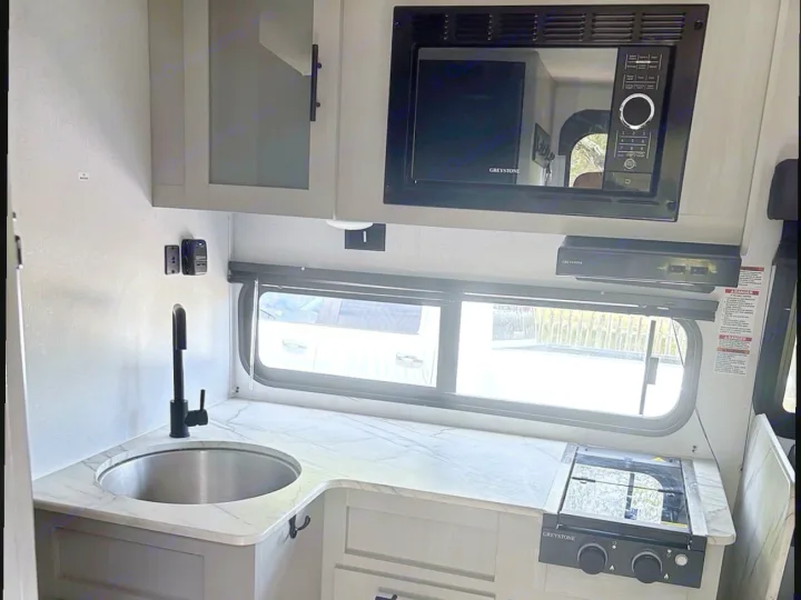 Modern RV rental kitchen interior with a sink, microwave, and stove, showcasing light grey cabinetry and marbled countertops.. Forest River Wildwood FSX 2022