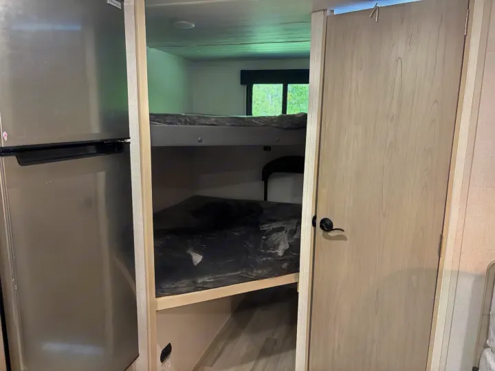 Interior of an RV rental showing a stainless steel refrigerator, two-level bunk beds with blankets, and a wooden door.. Keystone RV Springdale 2024