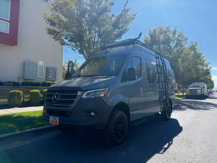 Gray Mercedes Sprinter camper van (an RV rental) with a roof rack and ladder, parked on a sunny street.. Jayco Terrain 2023