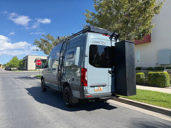 This gray Jayco camper van, an RV rental, features a roof rack, side ladder, and rear storage box on a sunny street.. Jayco Terrain 2023