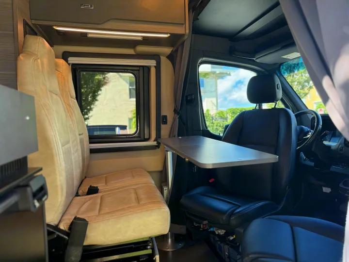 RV rental interior: A versatile dinette space with a swivel front passenger seat, table, and cream bench seating by the window.. Jayco Terrain 2023