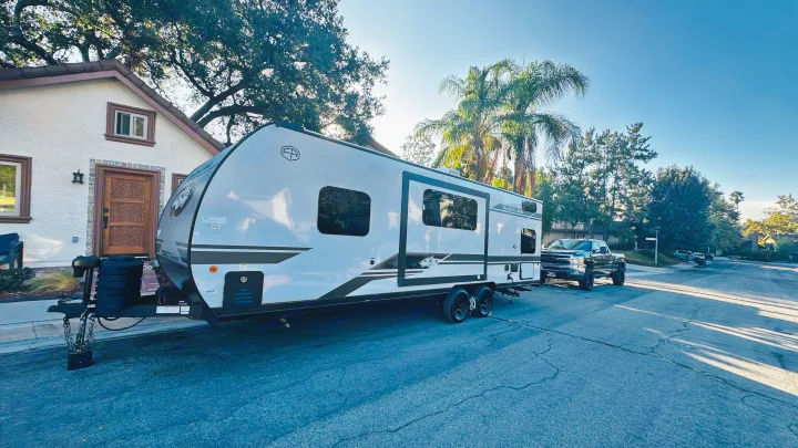 A modern travel trailer, ready for rv rental, is hitched to a pickup truck on a sunny residential street with a house & trees.. Forest River Cherokee Grey Wolf 2027