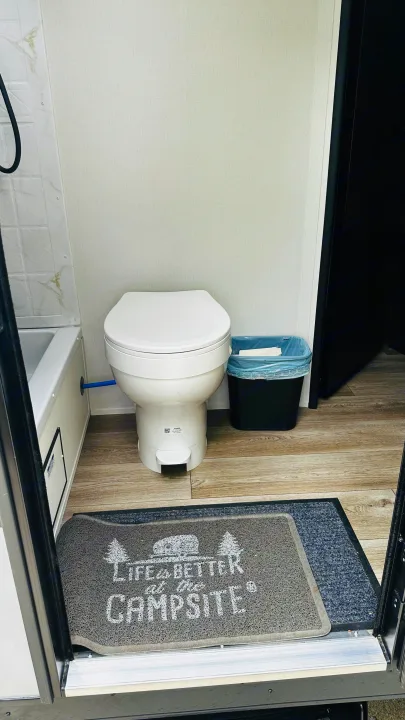 Inside an RV rental, this compact bathroom features a white foot-pedal toilet, shower area, bin, wood-look floor, and a welcome mat.. Forest River Cherokee Grey Wolf 2027