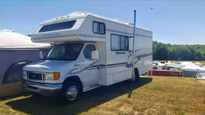 White Winnebago Minnie Class C RV rental, parked on grass at a sunny outdoor event with other tents & a plane.. Winnebago Minnie Winnie 2004