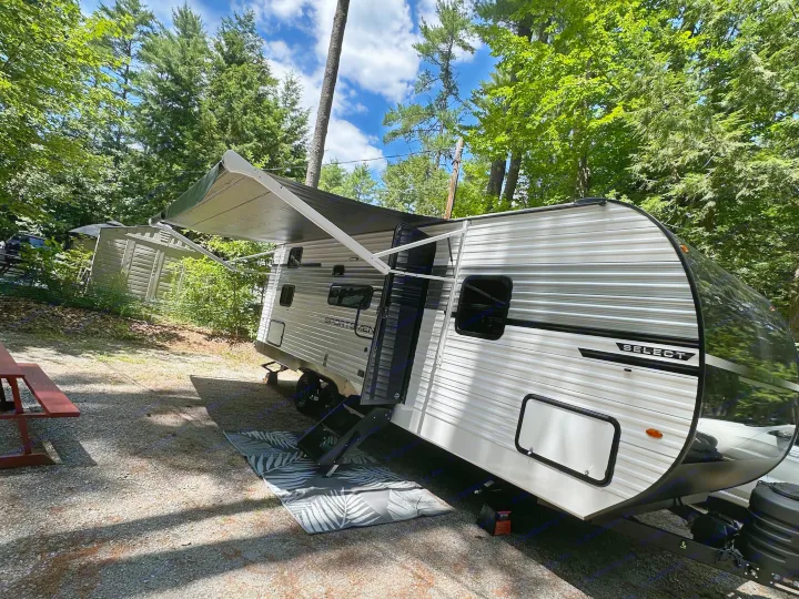 RV rental parked at a shaded campsite with an extended awning, picnic table, and forest backdrop under a blue sky.. KZ Sportsmen Select 2026