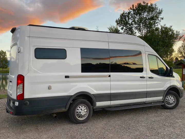 White camper van rental with sunset clouds reflecting in its windows, parked by water & trees on a gravel lot.. Ford Transit 2023