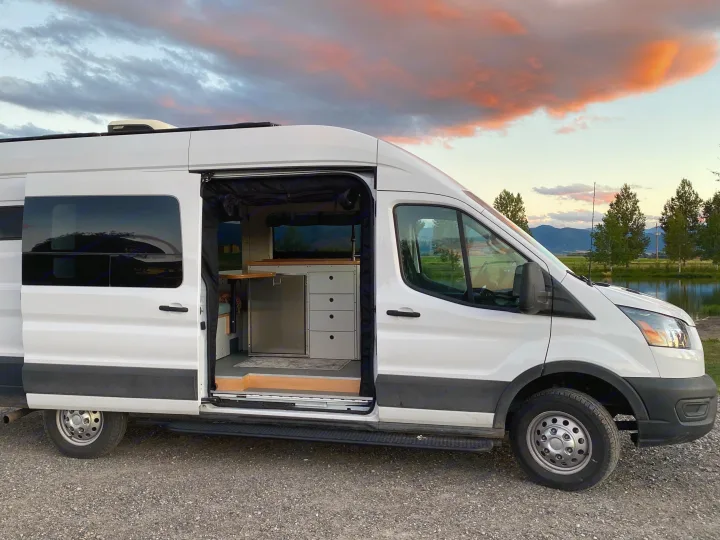 RV rental: White campervan with open door revealing its modern interior, parked lakeside at sunset. Ideal for van life adventures!. Ford Transit 2023