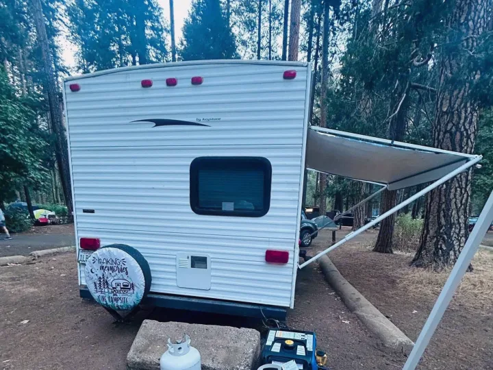 A white RV rental travel trailer with an extended awning and spare tire is parked in a treed campsite. Propane tank and generator visible.. Keystone RV Hideout 2009