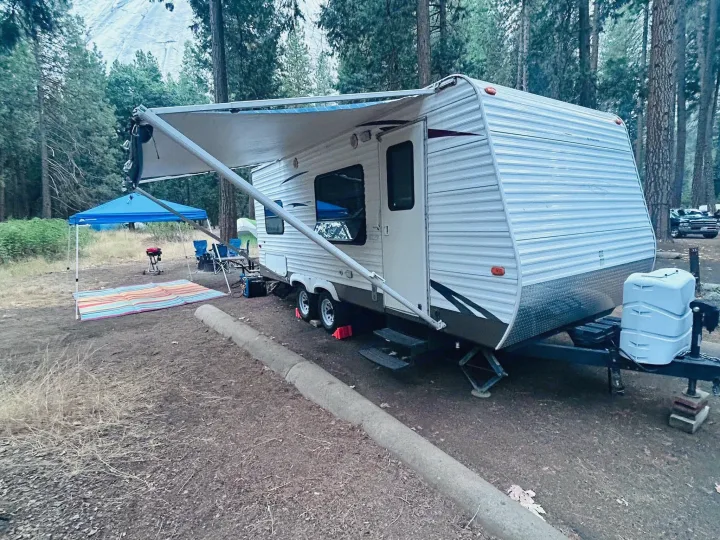 RV rental travel trailer parked in a wooded campsite with an open awning, blue pop-up tent, and a distant mountain peak.. Keystone RV Hideout 2009