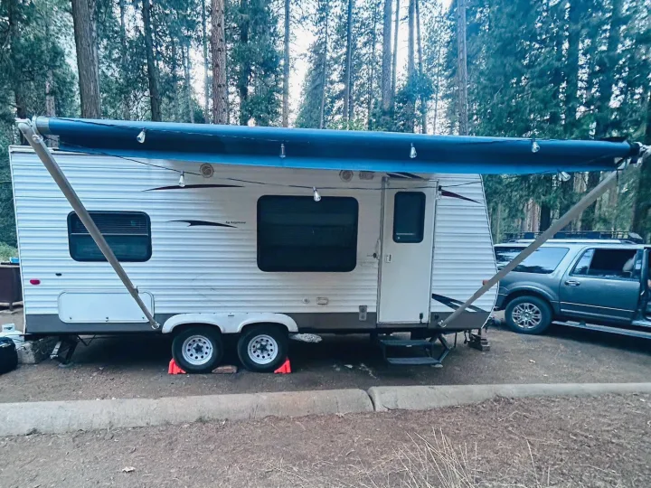 An RV rental, a white travel trailer, is set up at a shaded campsite with its blue awning extended & strung with lights.. Keystone RV Hideout 2009