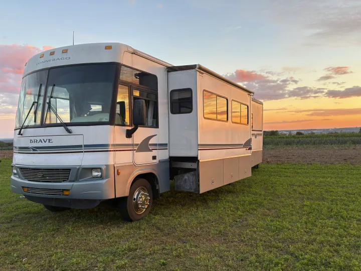This Winnebago Brave RV rental is parked on green grass, illuminated by the warm light of a sunset with pink-hued clouds.. Winnebago Brave 2004