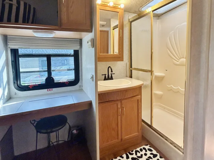 RV rental interior featuring a compact bathroom with shower and sink, plus a small desk area with a window.. Winnebago Brave 2004