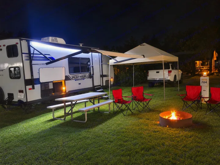 A bright RV rental campsite at night, featuring an illuminated RV, warm campfire, picnic table, and red chairs for a cozy outdoor escape.. Forest River Salem FSX 2019