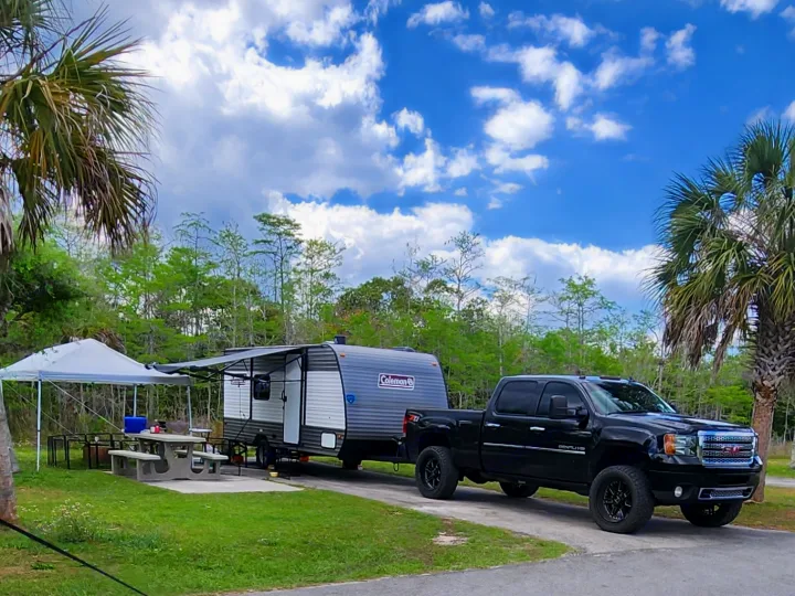 An RV rental, a travel trailer, is parked with a black pickup truck at a sunny campground. A picnic area with a canopy is set up.. Coleman R17 2024
