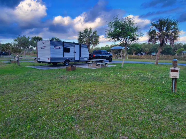 Coleman rv rental parked at a scenic campground with a pickup truck, picnic table, and fire pit under a cloudy sky.. Coleman R17 2024