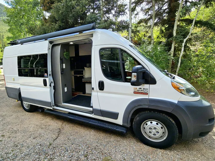 White camper van RV rental parked on a gravel road. Its side door is open, revealing a cozy interior, set against a lush forest.. Dodge Ram ProMaster 3500 2018