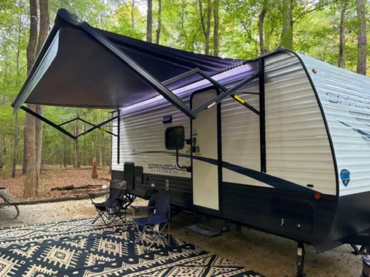 This inviting RV rental is perfectly set up at a wooded campsite, featuring an extended awning, outdoor chairs, and a patterned rug for comfortable outdoor living.. Keystone RV Springdale 2024