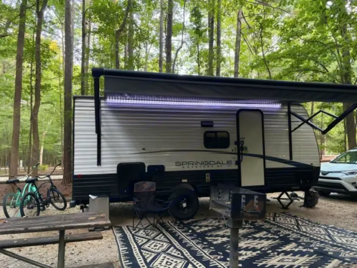 RV rental parked at a lush campground with an extended awning, bikes, and a picnic table. Perfect for a nature escape.. Keystone RV Springdale 2024