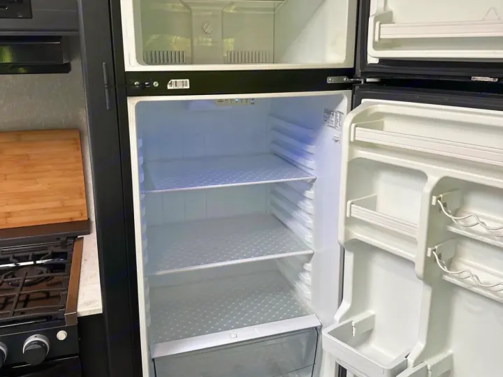 An RV rental's empty refrigerator and freezer are open, revealing clean shelves and door storage, beside a stove.. Forest River Cherokee 2022