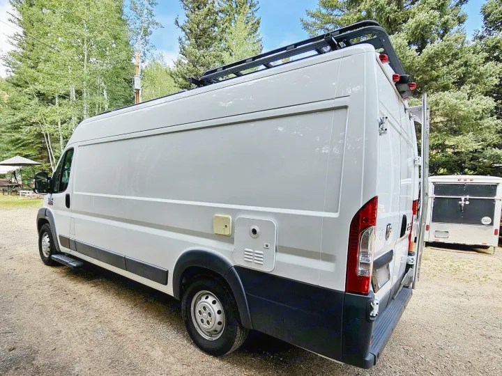 This white camper van RV rental, equipped with a roof rack, is parked on gravel amidst green trees.. Dodge Ram ProMaster 3500 2018