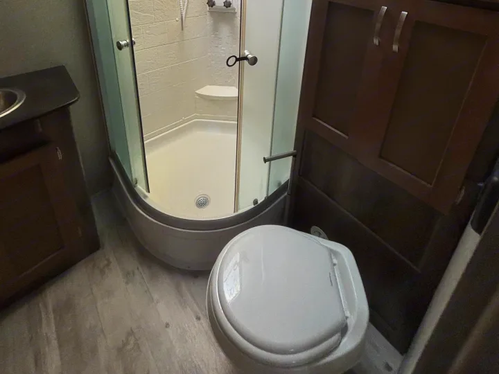 Compact RV rental bathroom featuring a corner shower, toilet, and sink vanity with dark wood cabinets.. Keystone RV Sprinter Campfire Edition 2018